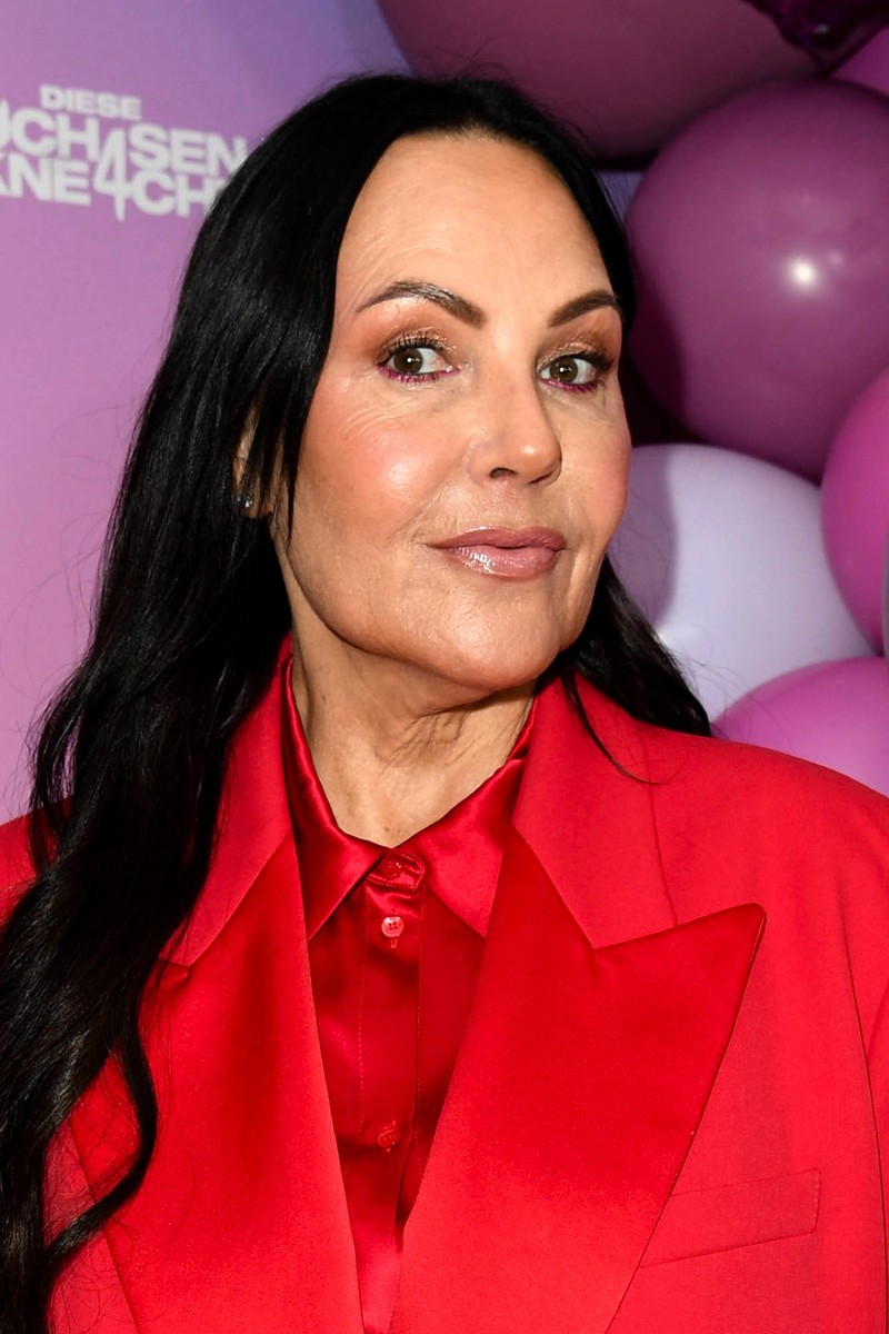 Auf dem Bild ist eine Person mit langen, schwarzen Haaren und einem roten Anzug zu sehen, die vor einer bunten Hintergrundkulisse posiert.